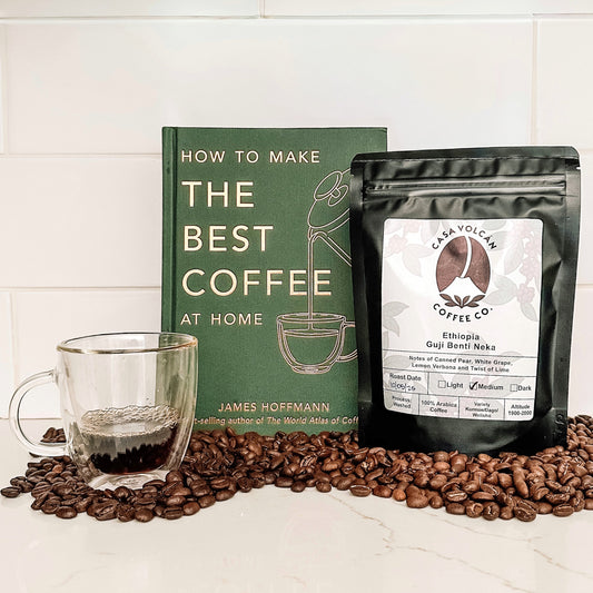 Coffee beans, a glass of coffee, a book on making coffee, and a coffee package on a white surface.