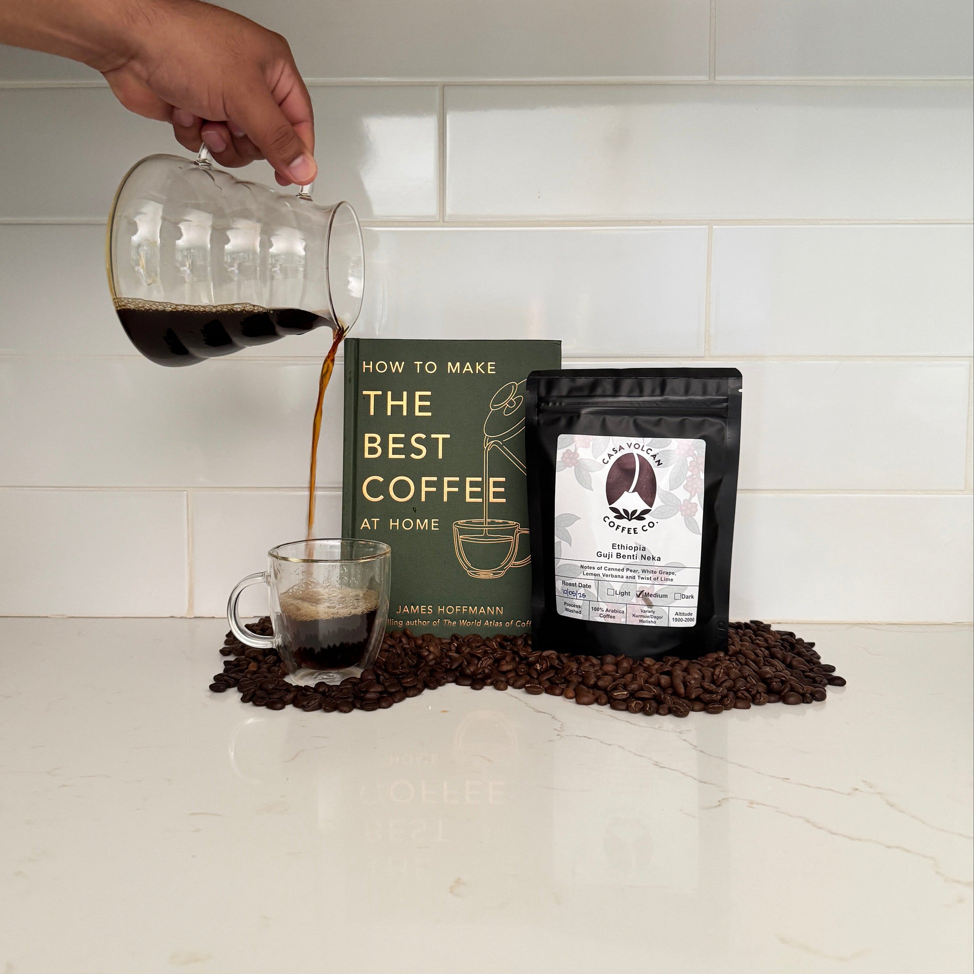 Person making coffee using a pour-over method with a book titled 'The Best Coffee' on a tiled wall.