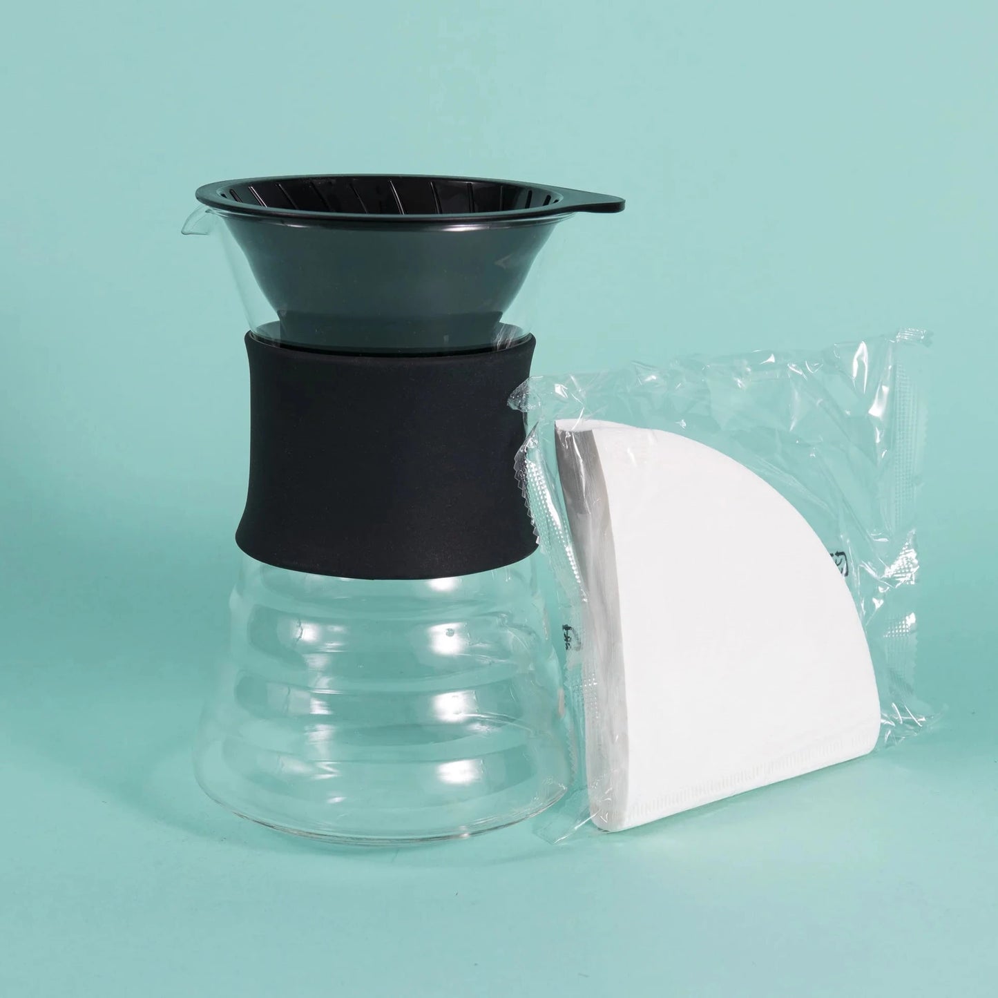 Clear glass coffee maker with black lid and white filter on a teal background